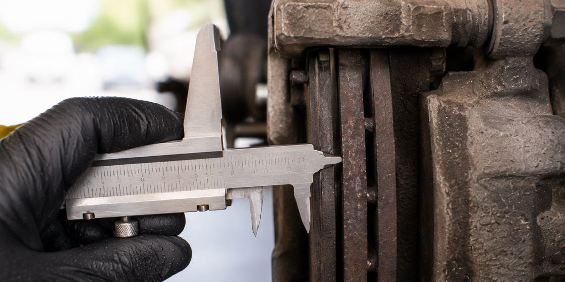 A mechanic measuring brake pad thickness with a vernier caliper at the wheel of a car on a lift